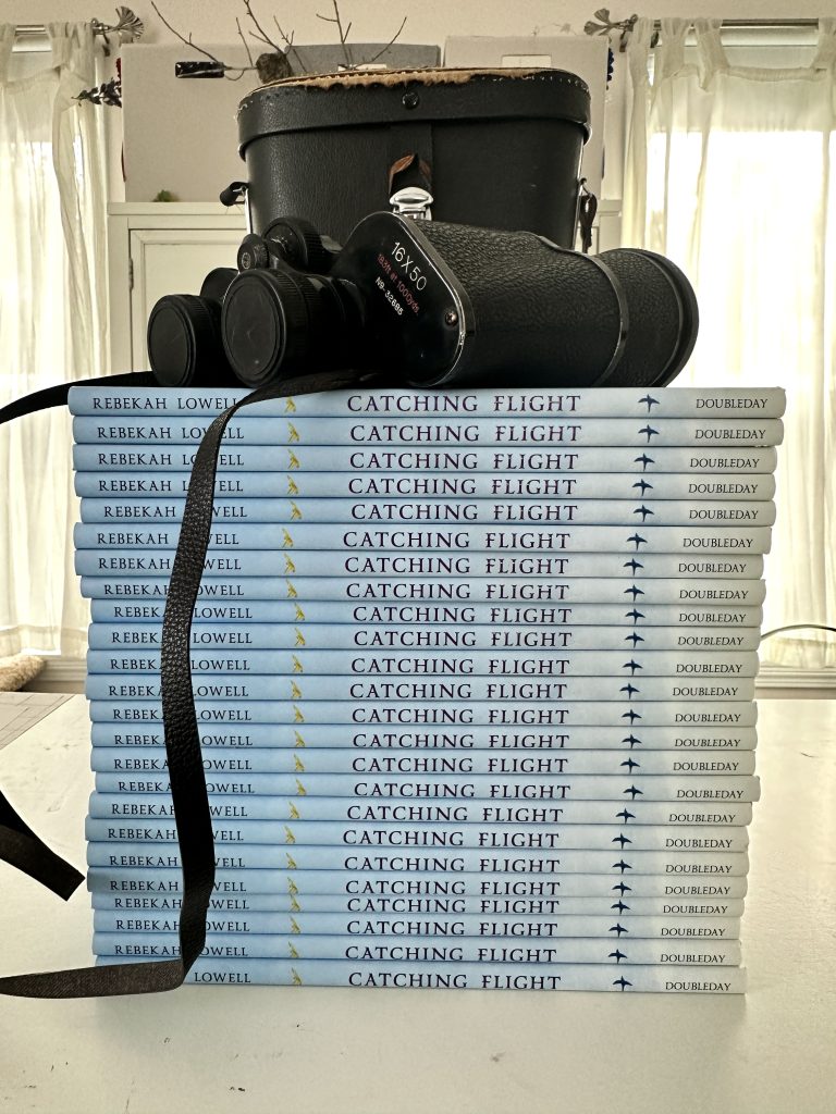 A stack of Catching Flight books by Rebekah Lowell on a table with a pair of bird-watching binoculars on top.