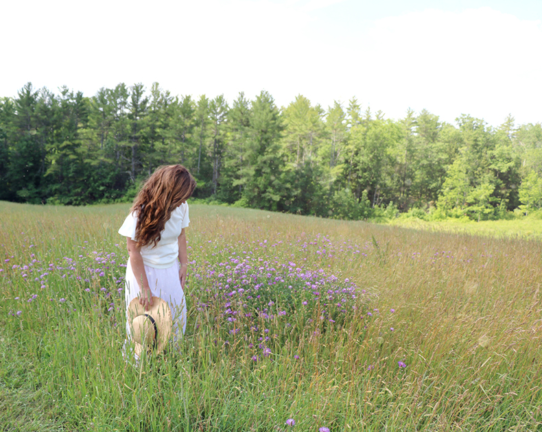 Author/illustrator, Rebekah Lowell in a field of wildflowers, looking down and holding a wide-brimmed hat.