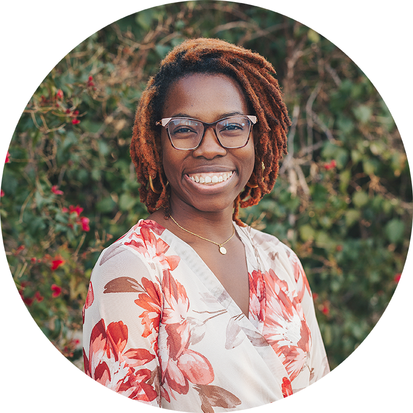 Illustrator and author, Acamy Schleikorn smiles in an outdoor garden backdrop. Academy is a Black woman with glasses, hair in locs, and in this photo wears a floral blouse.