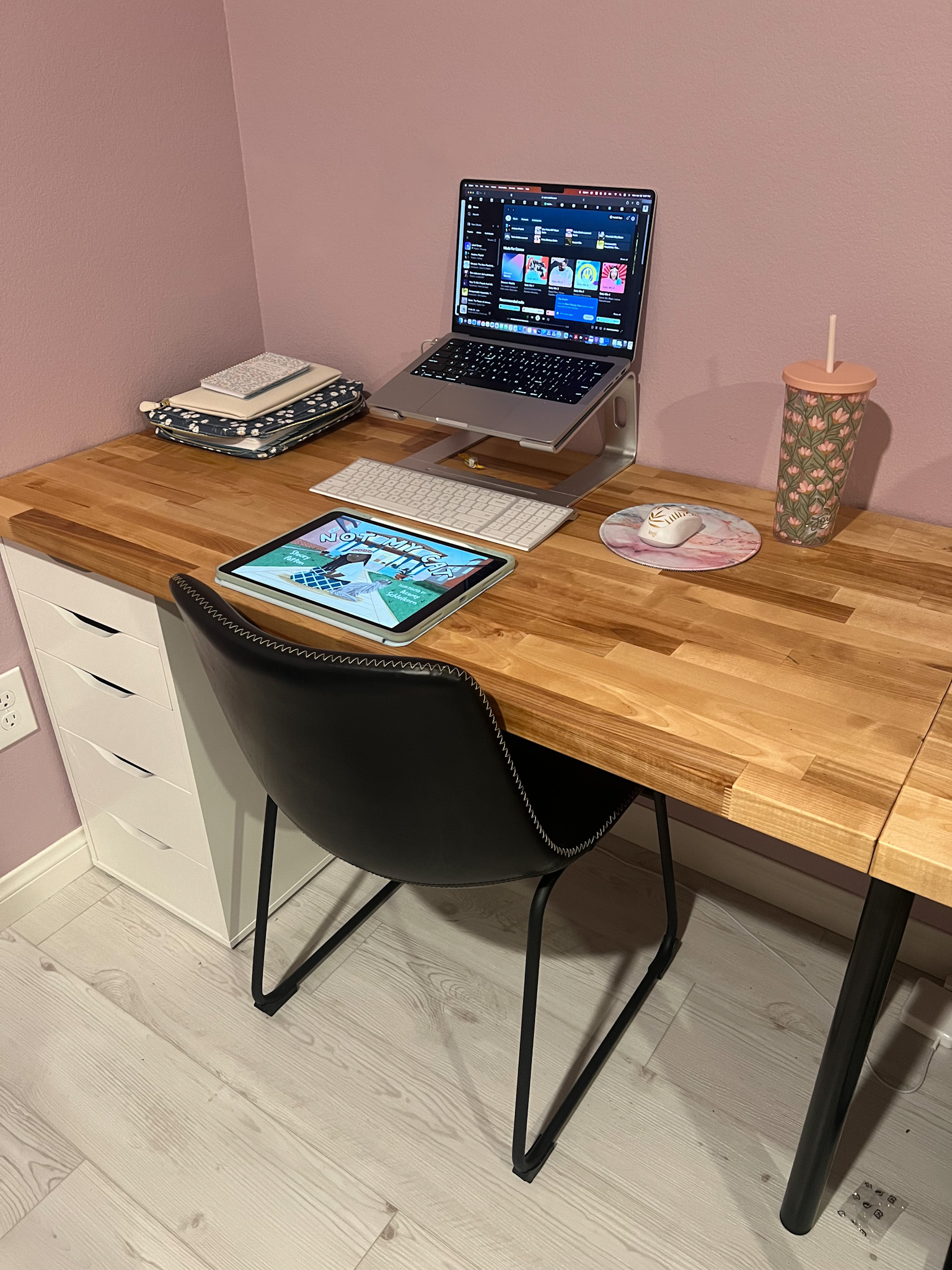 A wooden desk with an iPad and MacBook on top with a black chair and some white filing drawers below.