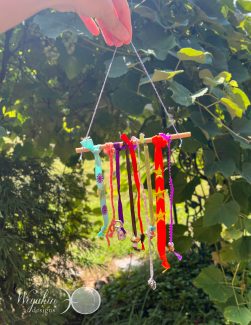 A fairy charm made from a wood stick, ribbons, yarn, string, beads, and sequins sparkling in the sun in front of some leaves.