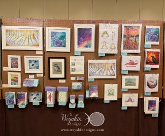 A display of Victoria's art on a brown peg board panel at Arisia in January of 2024.