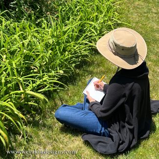 A person sits in the grass next to some plants while drawing in a sketch book.