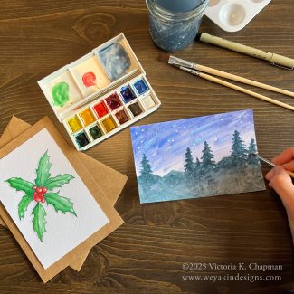 A table set up with watercolors, paint brushes, water, and mixing pans. One painting of holly leaves and berries is mounted on a card with an envelope. The other painting is in-progress being painted with trees and a night sky. ©2025 Victoria K. Chapman.