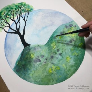 A hand with a paintbrush, paints a scene of a green spring tree with dark contrasted bark and green, flowery grass in watercolors.