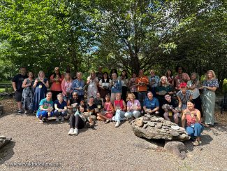 A group of various authors and illustrators stand in a gravel area outdoors under trees in the Boyds Mills “word garden". Each person is holding a stone with a word on it.