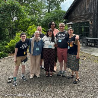 A small group of people stand together smiling outside next to a grey/brown barn with trees in the background.